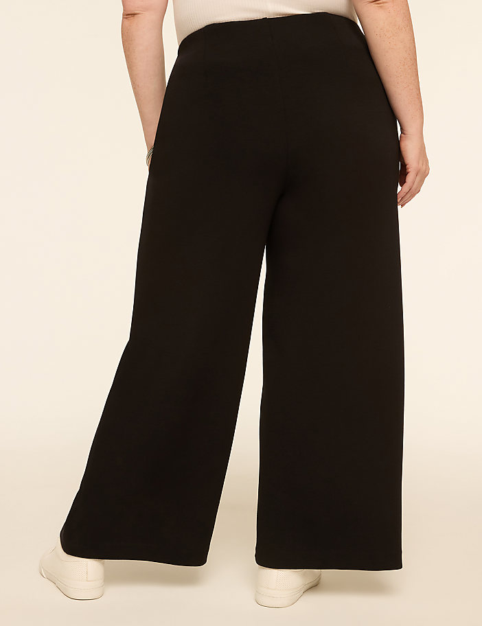 A person wearing black wide-leg pants and white sneakers stands against a plain, light-colored background.