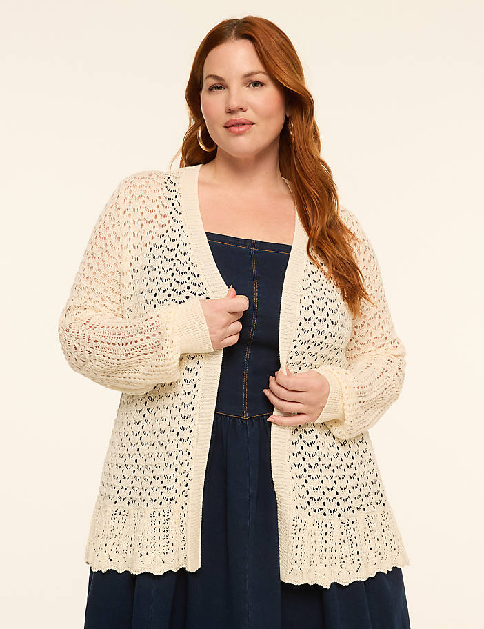 A woman with red hair stands in a cream-colored, open-knit cardigan over a dark denim dress against a plain, light background.