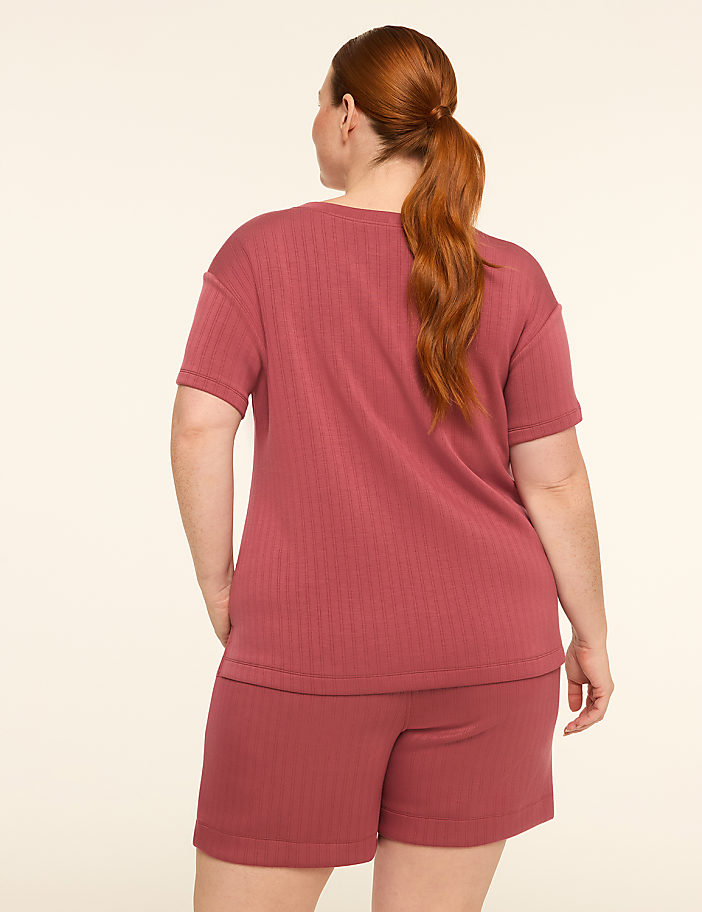 A woman with auburn hair pulled into a ponytail stands with her back to the camera, wearing a ribbed rose-colored t-shirt and matching shorts against a plain, light-colored background.