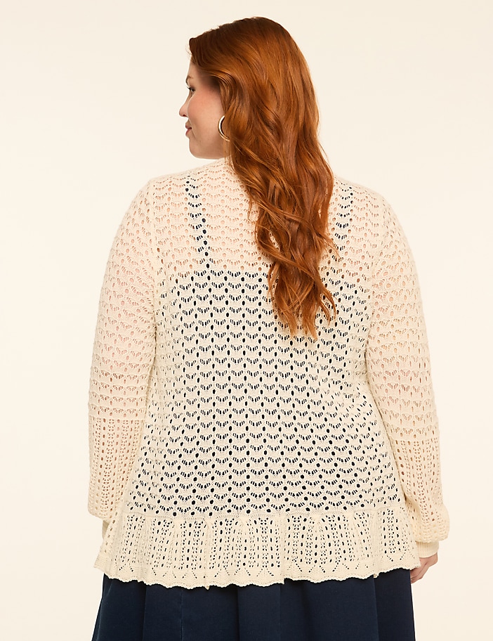 A woman with red hair wearing a cream-colored crocheted cardigan stands against a plain, light-colored background.