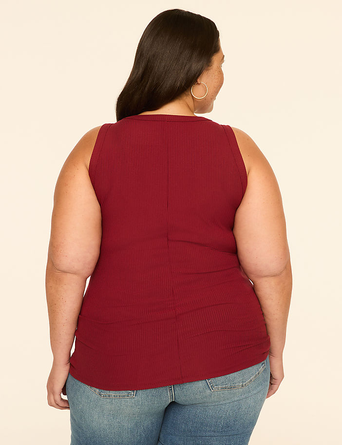 A woman in a maroon ribbed tank top and blue jeans stands against a plain cream background.