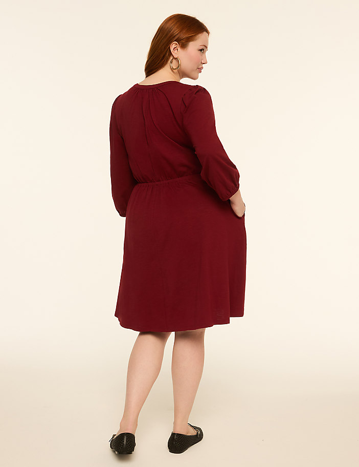 A woman in a maroon dress with her back to the camera stands against a plain, light-colored background.
