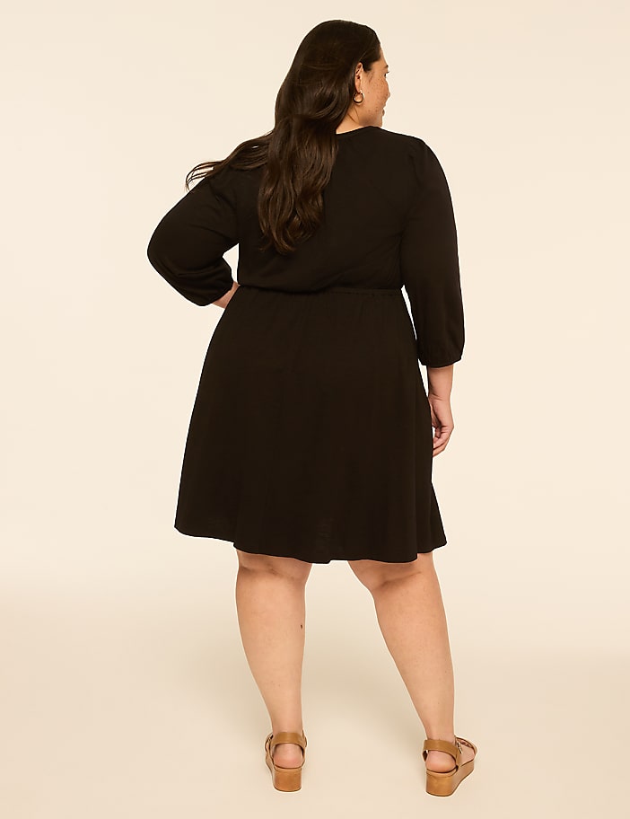 A woman in a black dress stands with her back to the camera against a plain, light-colored background.