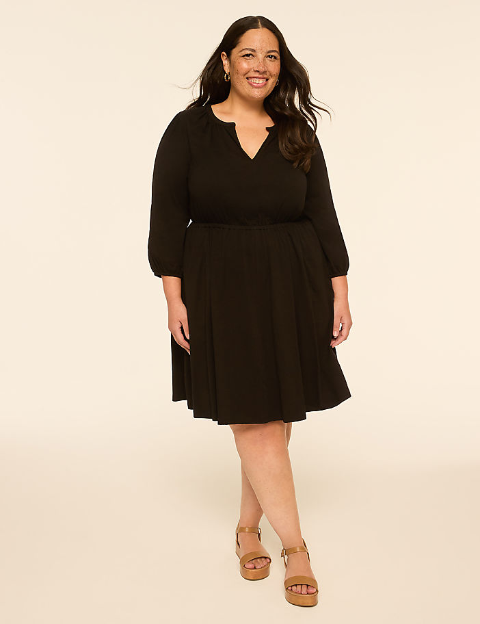 A smiling woman in a black dress and tan sandals stands against a plain, light-colored background.