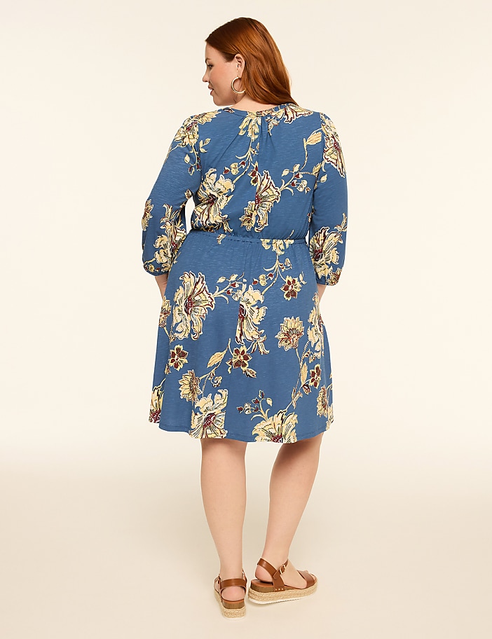 A woman in a blue floral dress stands with her back to the camera against a plain, light-colored background.