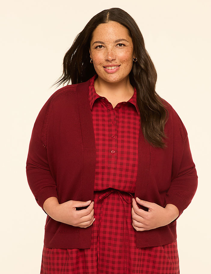 A smiling woman with dark hair and freckles, wearing a maroon cardigan over a red gingham dress, stands against a plain cream background.