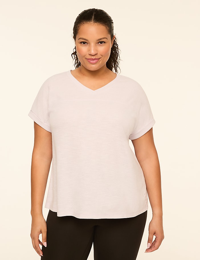 A woman with dark hair and a v-neck shirt stands against a plain, light-colored background.