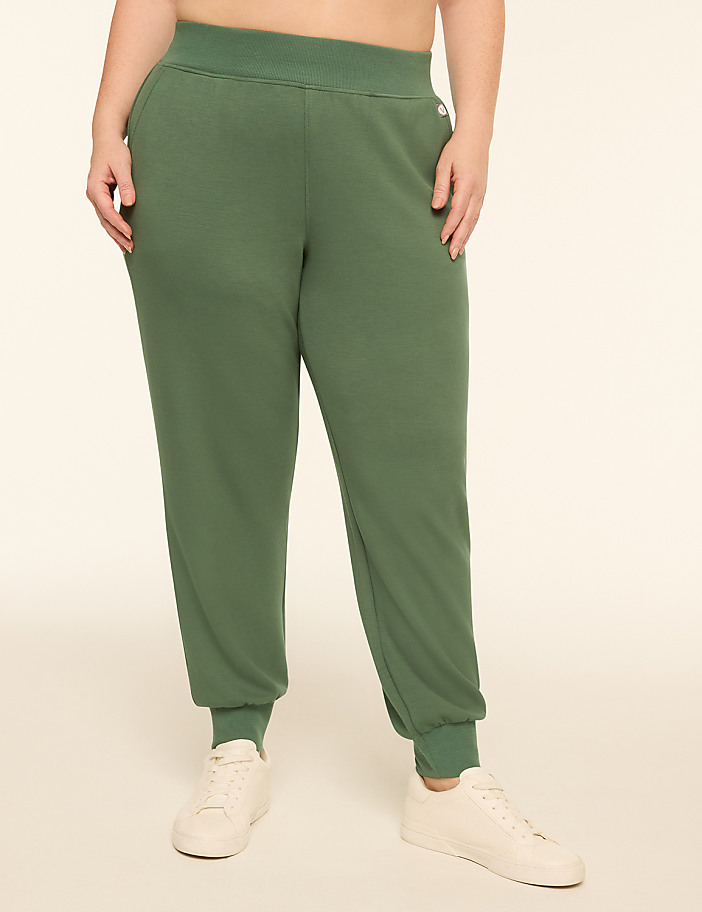 A person wearing green sweatpants and white sneakers stands against a plain, light-colored background.