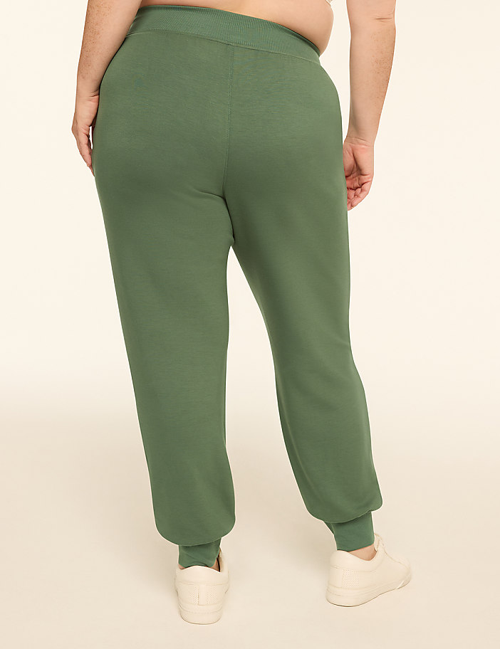 A person wearing green sweatpants and white sneakers stands against a plain, light-colored background.