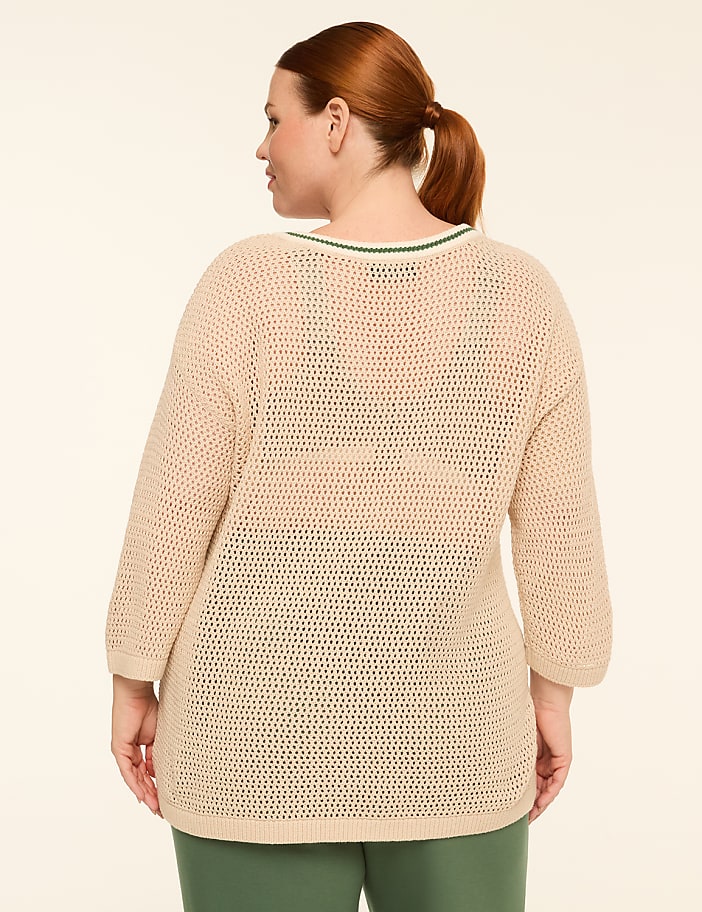 A woman with auburn hair pulled back in a ponytail stands facing away from the camera, wearing a loose-fitting, cream-colored, open-knit sweater with green trim at the neckline, and green pants, against a plain, light beige background.