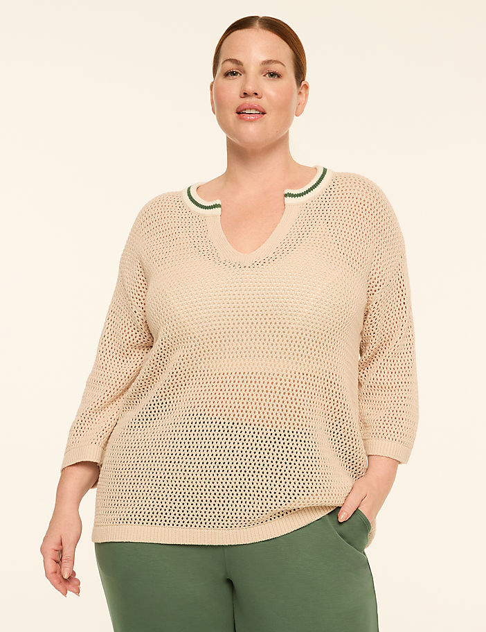 A woman in a beige, open-knit sweater with green trim stands against a plain, light-colored background.