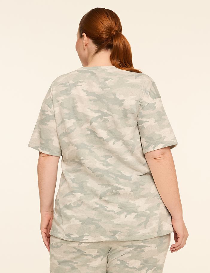 A woman with red hair pulled back in a ponytail stands facing away from the camera, wearing a camouflage t-shirt and pants against a plain, light-colored background.
