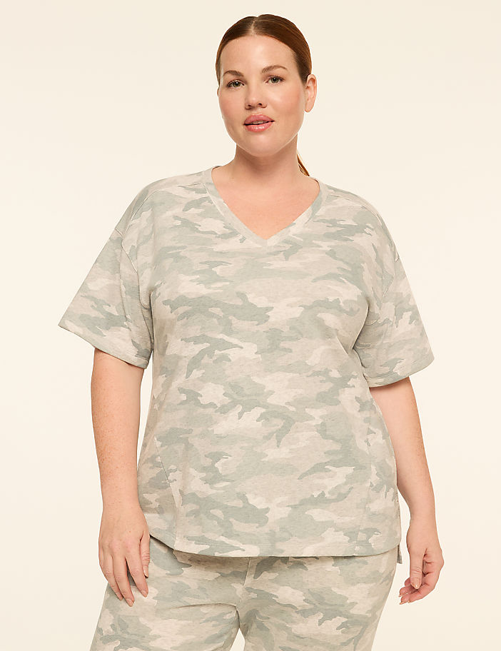 A woman in a light green camouflage v-neck t-shirt and matching pants stands against a plain, light beige background.