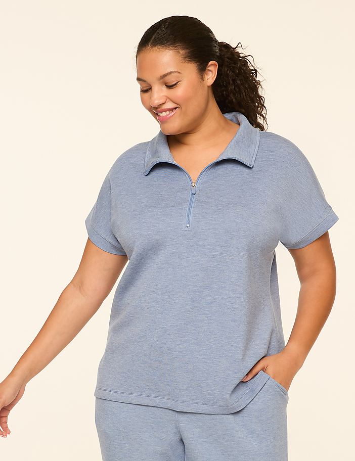 A smiling woman in a light blue zip-neck top and matching pants stands against a plain, light background.