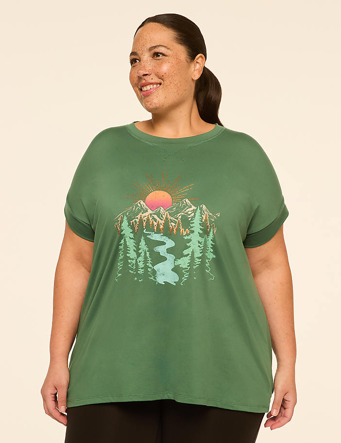 A woman in a green t-shirt with a mountain and river graphic stands against a plain background.
