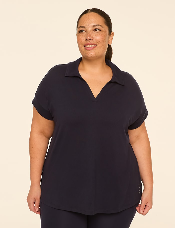 A smiling woman with freckles in a navy polo shirt stands against a plain, light beige background.