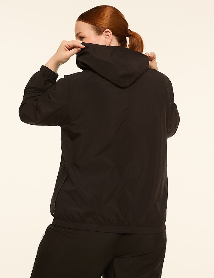 A woman in a black hooded jacket adjusts the hood against a plain, light-colored background.