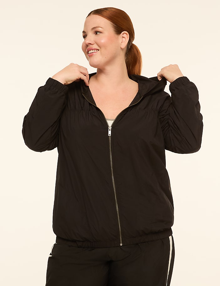 A smiling woman in a black hooded jacket stands against a plain, light-colored background.