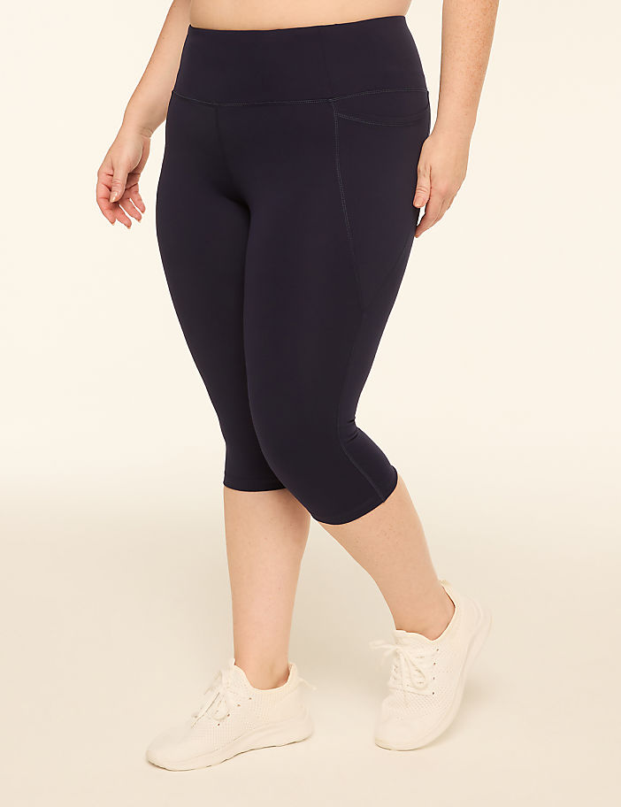 A woman in navy capri leggings and white sneakers stands against a plain, light-colored background.