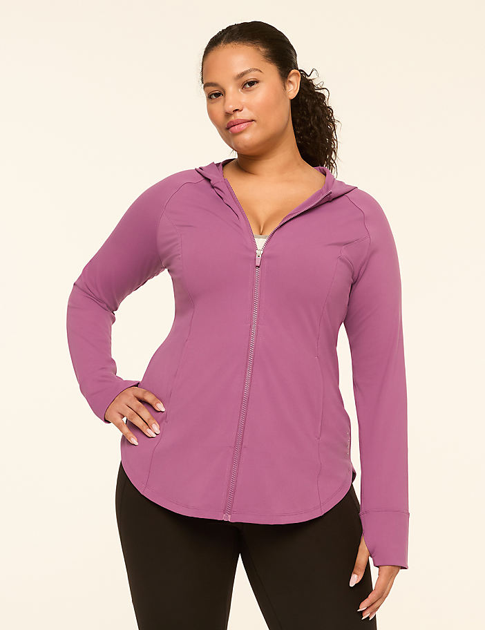 A woman in a purple zip-up hoodie and black leggings stands against a plain, light-colored background.