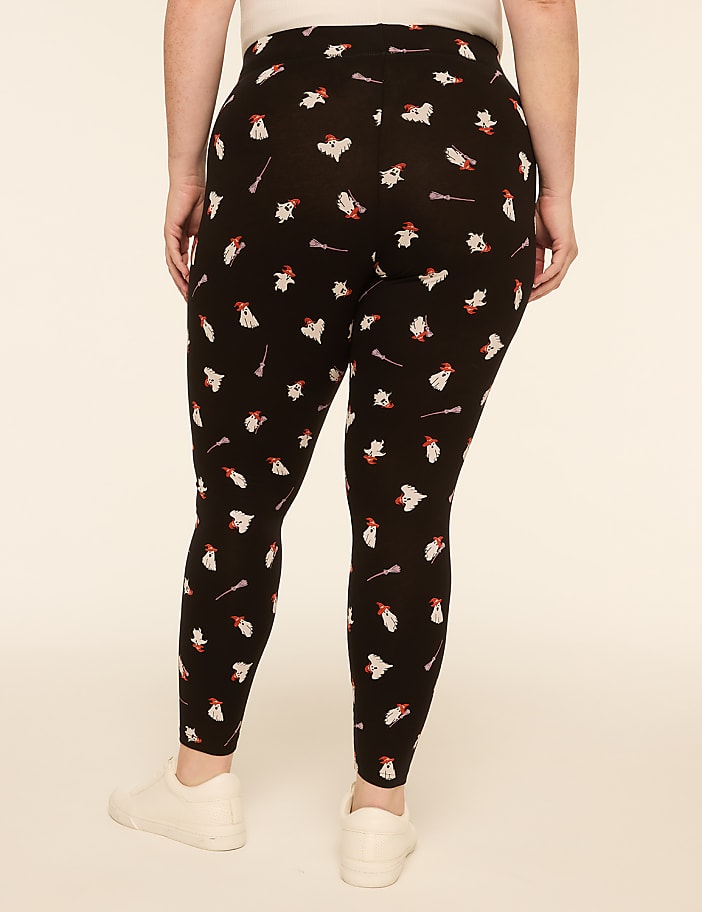 A person wearing black leggings with a repeating pattern of white ghosts and red candy canes stands against a plain, light beige background.