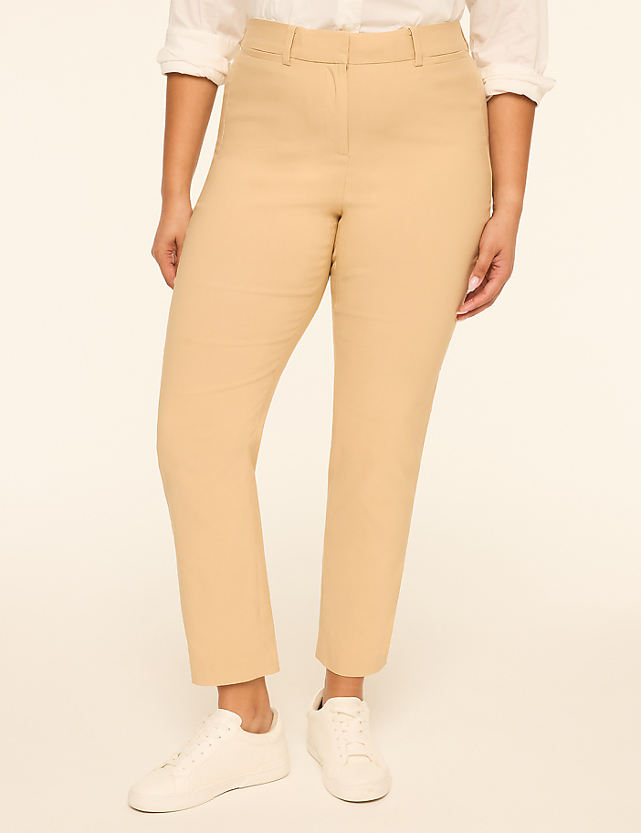A woman in a white shirt and tan pants stands against a plain, light background.
