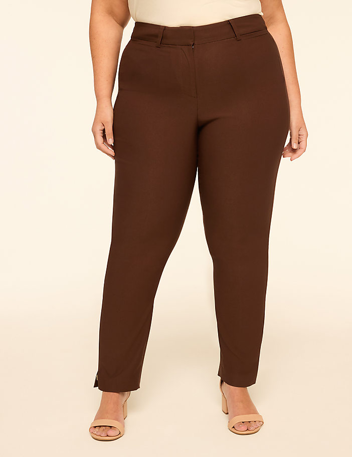 A woman in brown pants and a cream top stands against a plain cream background.