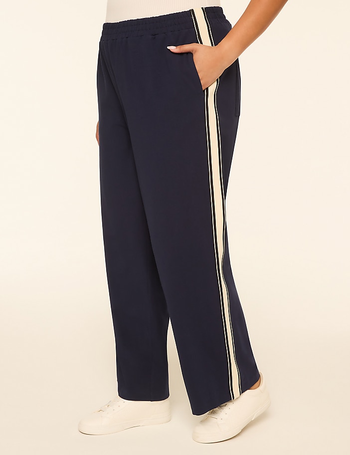 A person's legs are shown from the knee down, wearing navy blue pants with a cream stripe down the side and white sneakers, against a plain cream background.