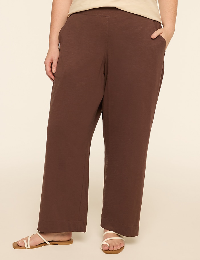A person wearing brown pants and white sandals stands against a plain, light-colored background.