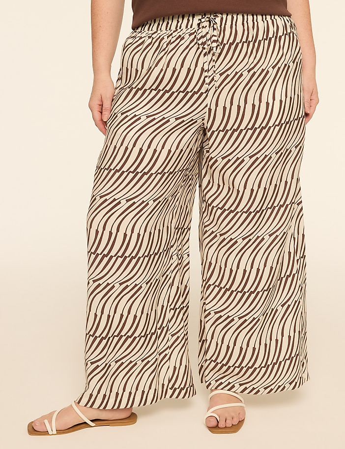 A person stands wearing wide-leg pants with a brown and cream wavy pattern, against a plain, light-colored background.