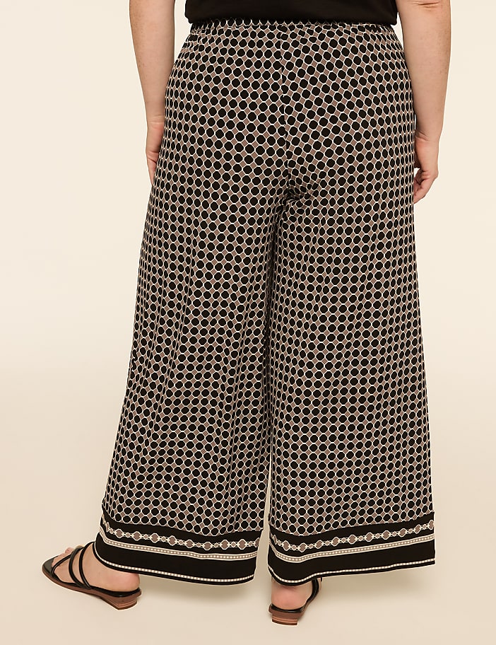 A person stands wearing patterned wide-leg pants and sandals against a plain, light-colored background.