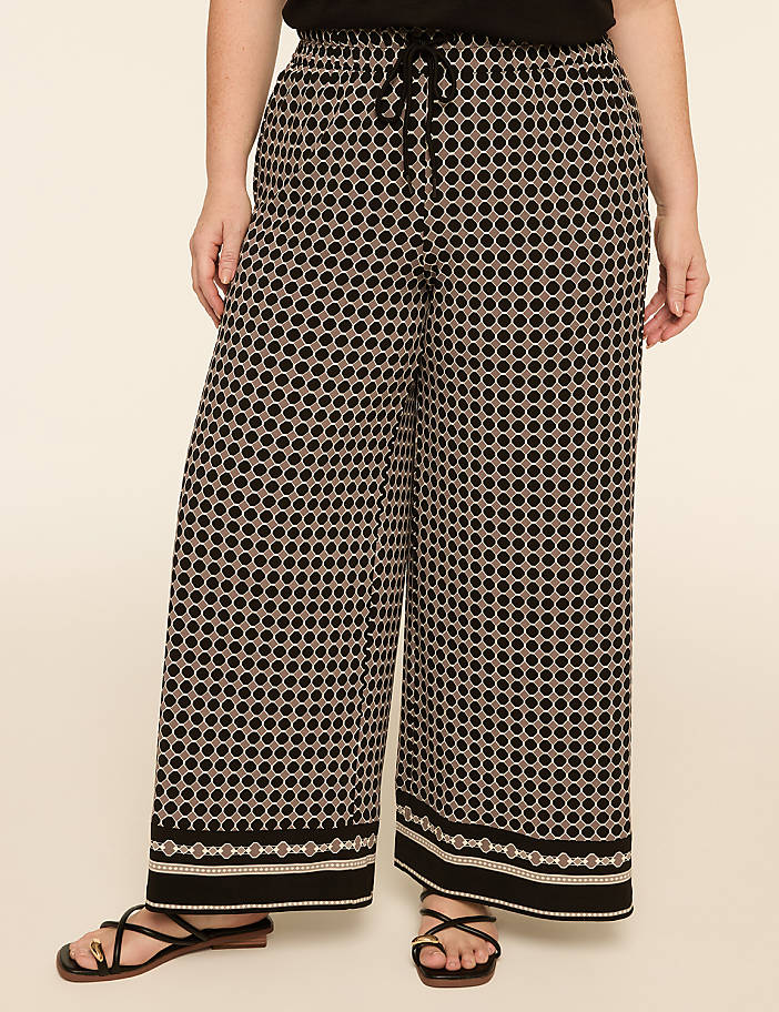 A person stands in wide-leg patterned pants with a geometric design and a plain background.