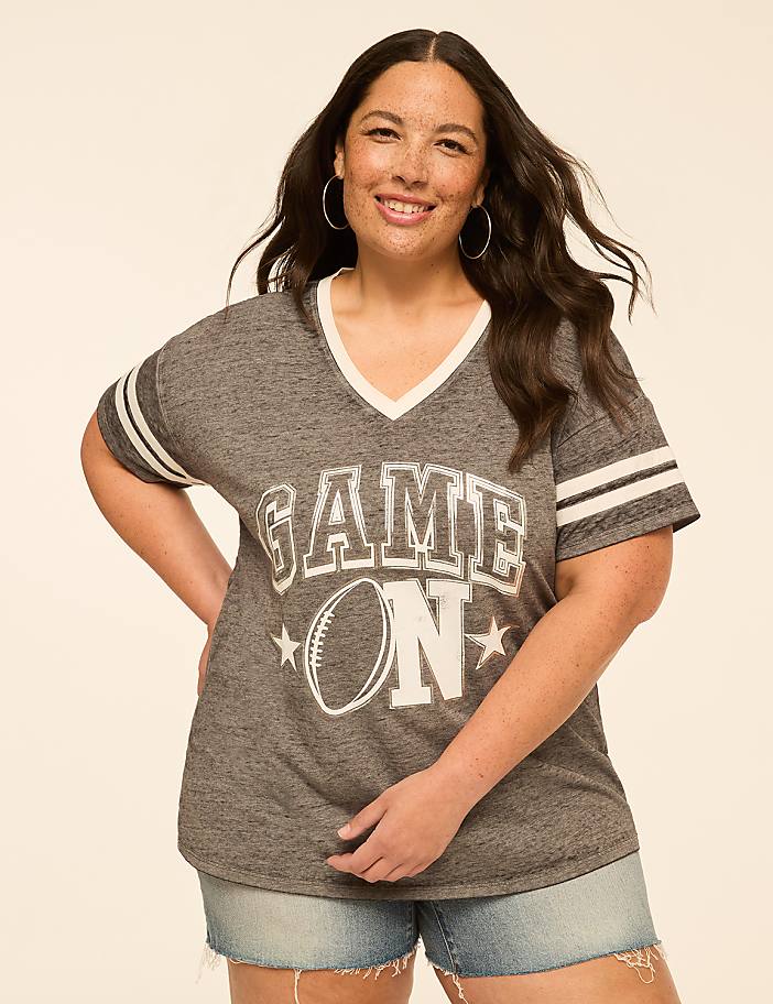 A smiling woman in a "GAME ON" t-shirt and denim shorts stands against a plain, light-colored background.