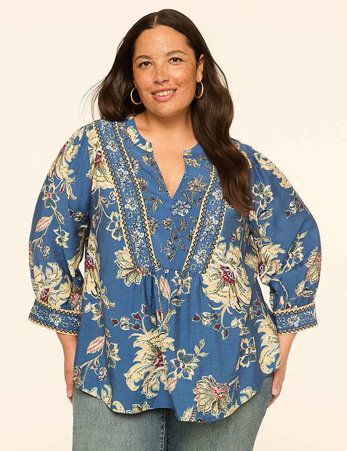 A woman in a blue floral blouse stands against a plain, light-colored background.