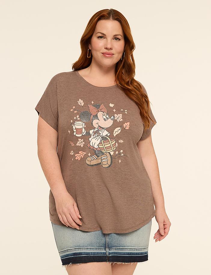 A woman with auburn hair and a denim skirt stands against a plain, light brown background, wearing a brown t-shirt with a graphic of Minnie Mouse holding a coffee cup and a basket of leaves.