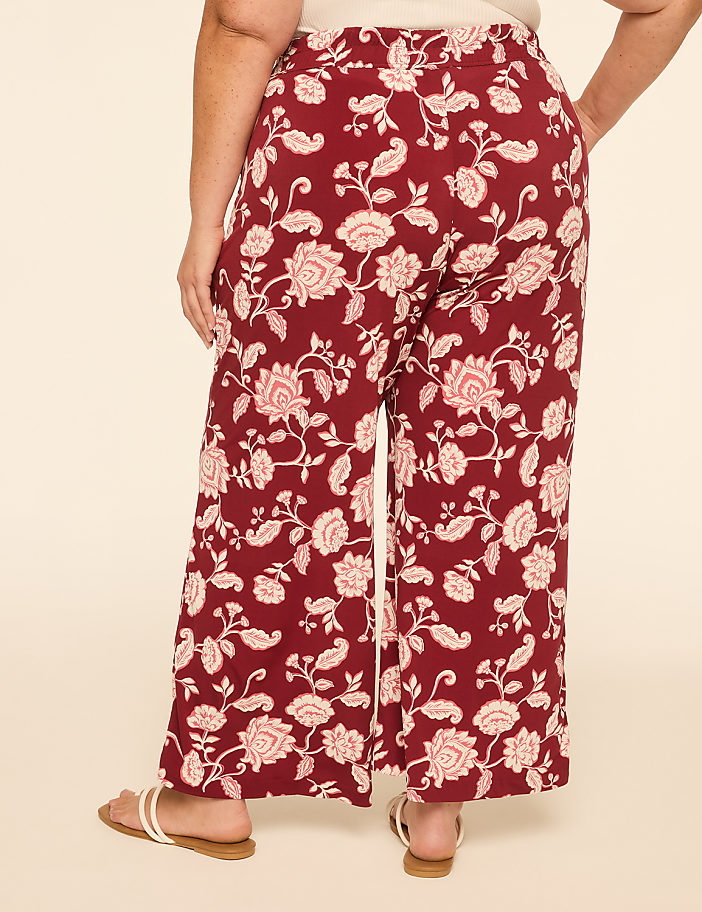 A person wearing maroon floral wide-leg pants and white sandals stands against a plain cream background.