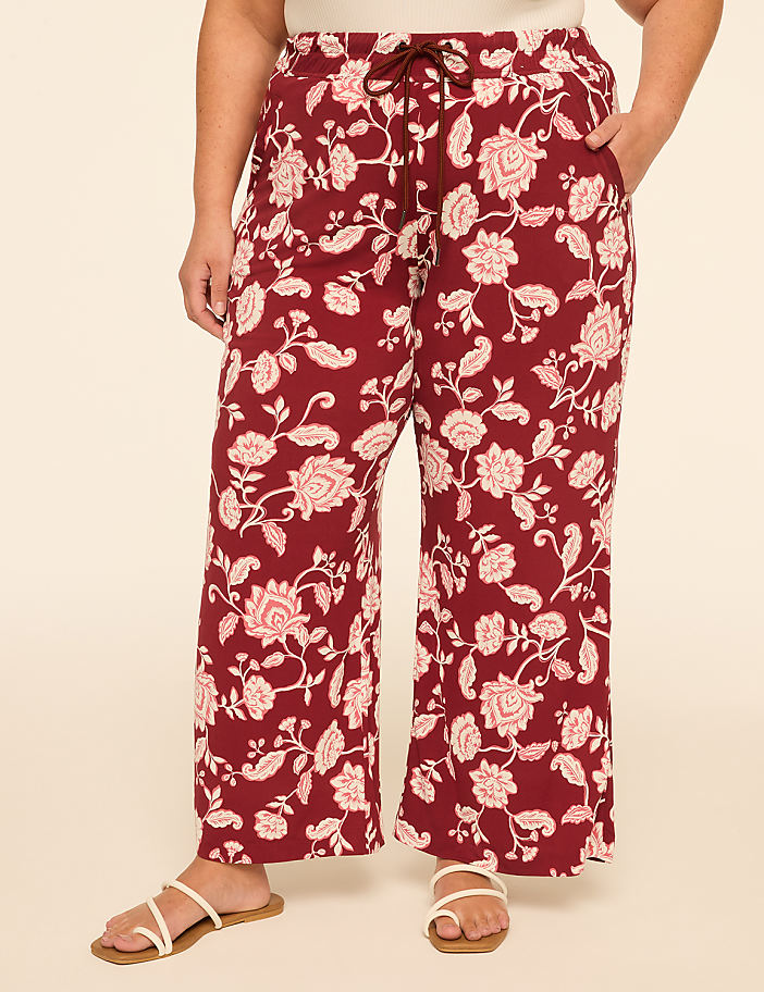 A person wearing maroon floral-patterned pants and white sandals stands against a plain beige background.
