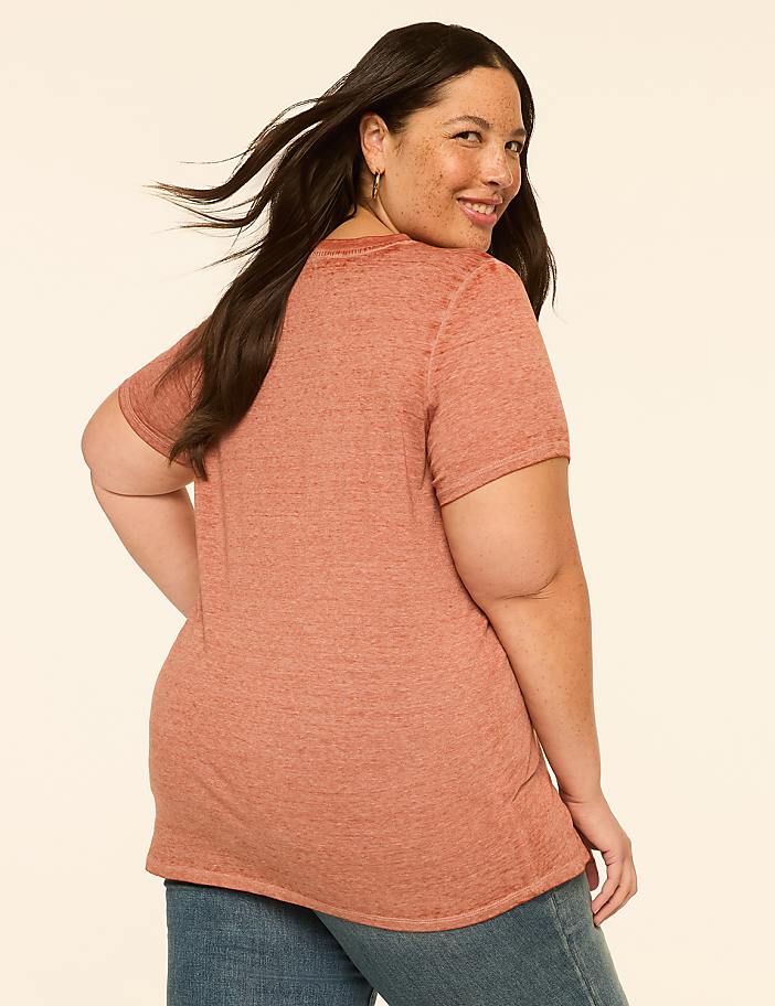 A woman in a rust-colored t-shirt and jeans smiles over her shoulder against a plain, light-colored background.