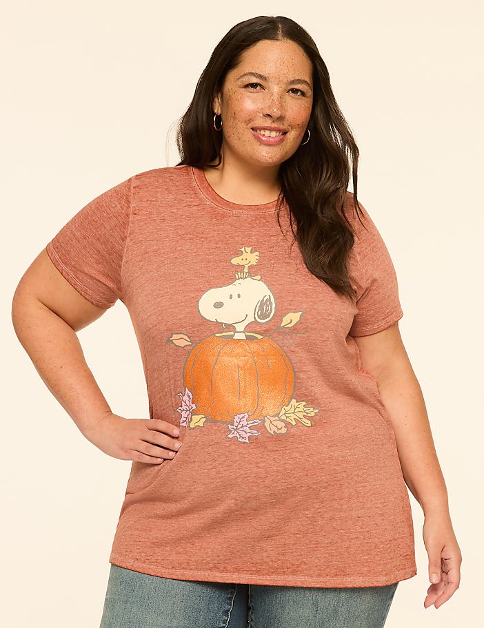 A woman in a rust-colored t-shirt with a Snoopy pumpkin graphic stands against a plain, light-colored background.