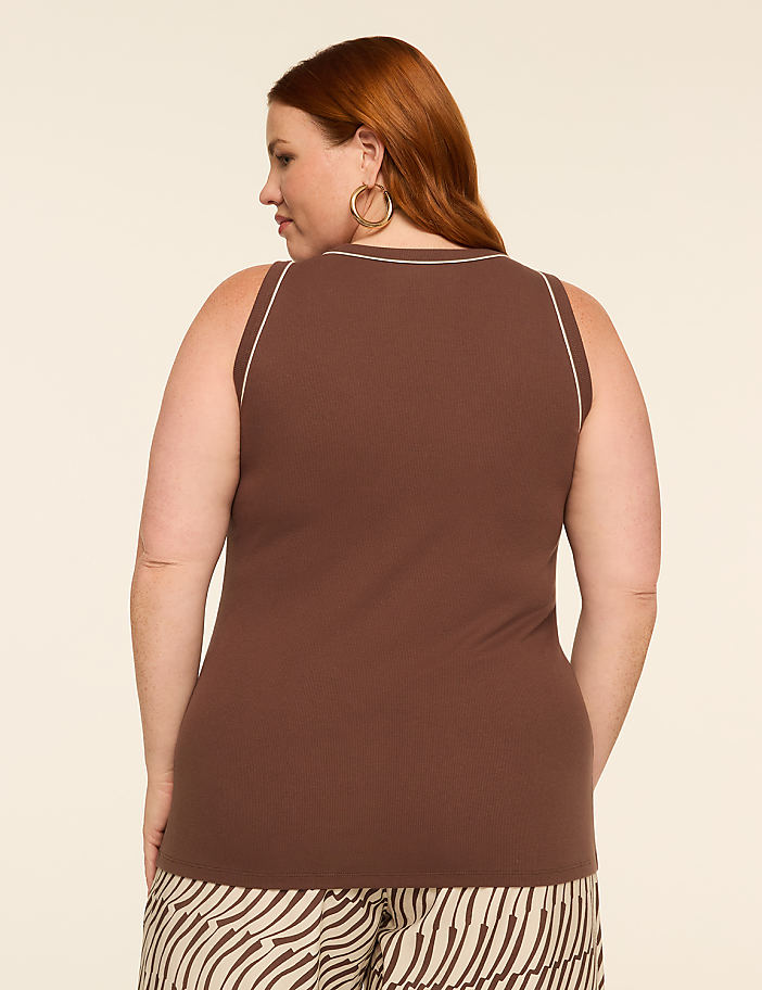 A woman with red hair, wearing a brown ribbed tank top with white trim and patterned pants, stands against a plain beige background.