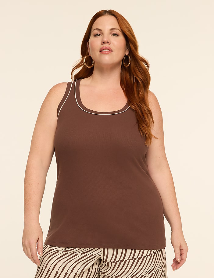 A woman with auburn hair and hoop earrings poses in a brown ribbed tank top with white trim, against a plain, light-colored background.
