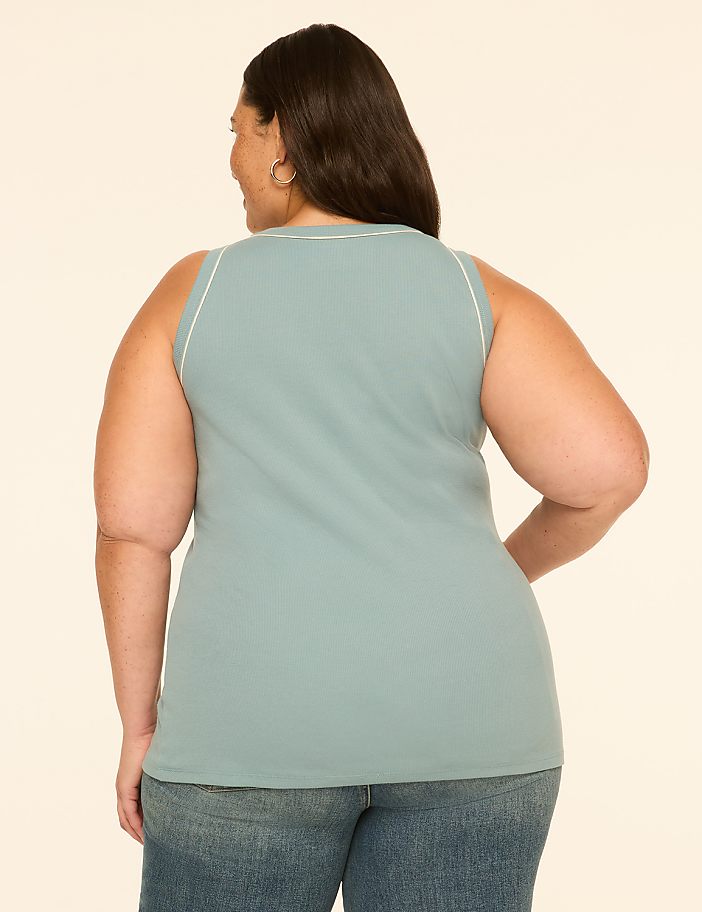 A woman in a teal ribbed tank top and blue jeans stands against a plain cream background.