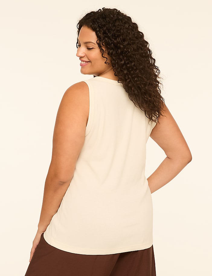 A woman in a cream-colored tank top and brown pants stands against a plain, light-colored background.