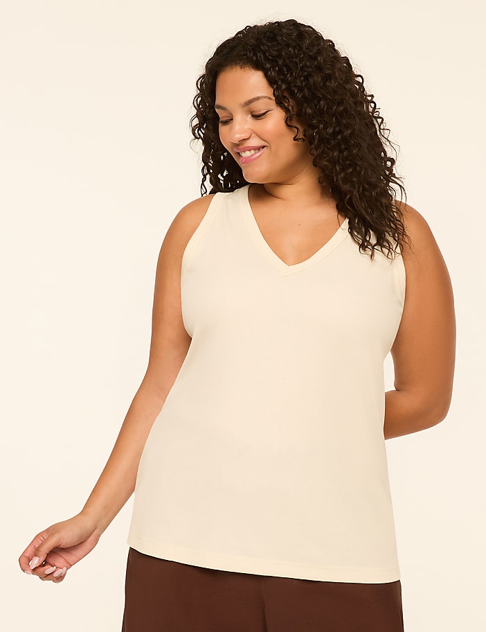 A smiling woman with curly dark hair wears a cream-colored v-neck tank top and brown pants against a plain, light background.