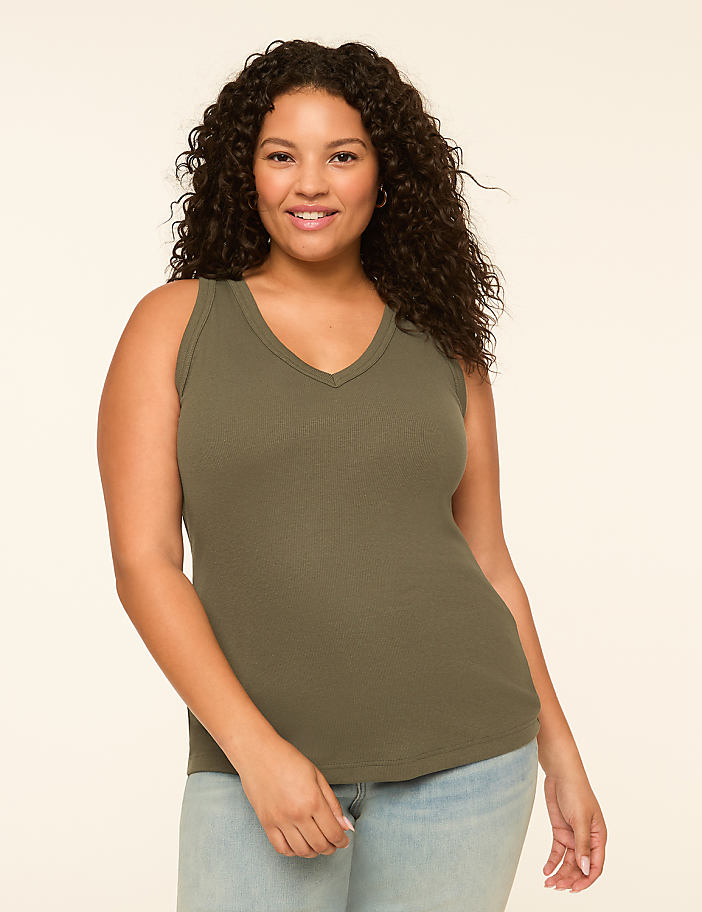 A smiling woman with curly dark hair wears a green ribbed tank top and light blue jeans against a plain cream background.