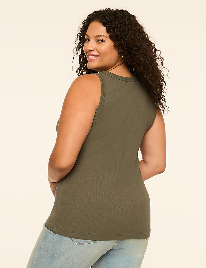 A woman in a green ribbed tank top and light blue jeans smiles over her shoulder against a plain cream background.
