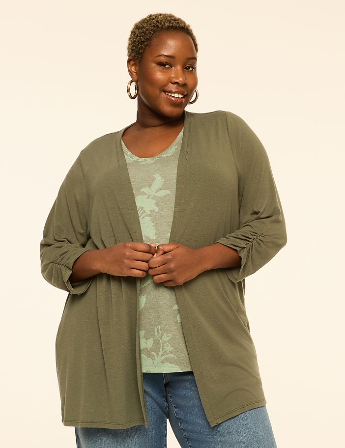 A smiling woman in a green cardigan and floral top stands against a plain, light background.
