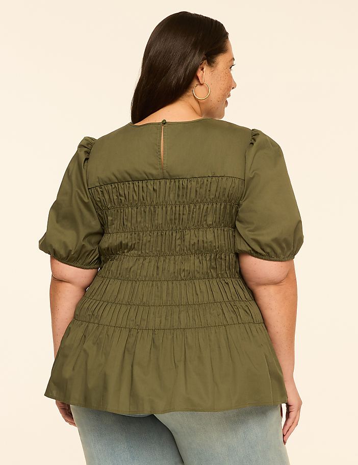 A woman in a green smocked top and light blue jeans stands against a plain beige background.