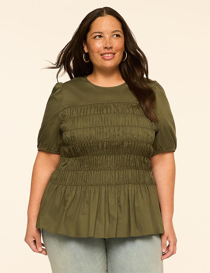 A smiling woman with dark hair and hoop earrings poses in a green smocked top and light blue jeans against a plain, light-colored background.