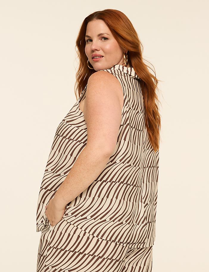 A woman with red hair and a patterned sleeveless top stands with her hands in her pockets against a plain, light-colored background.
